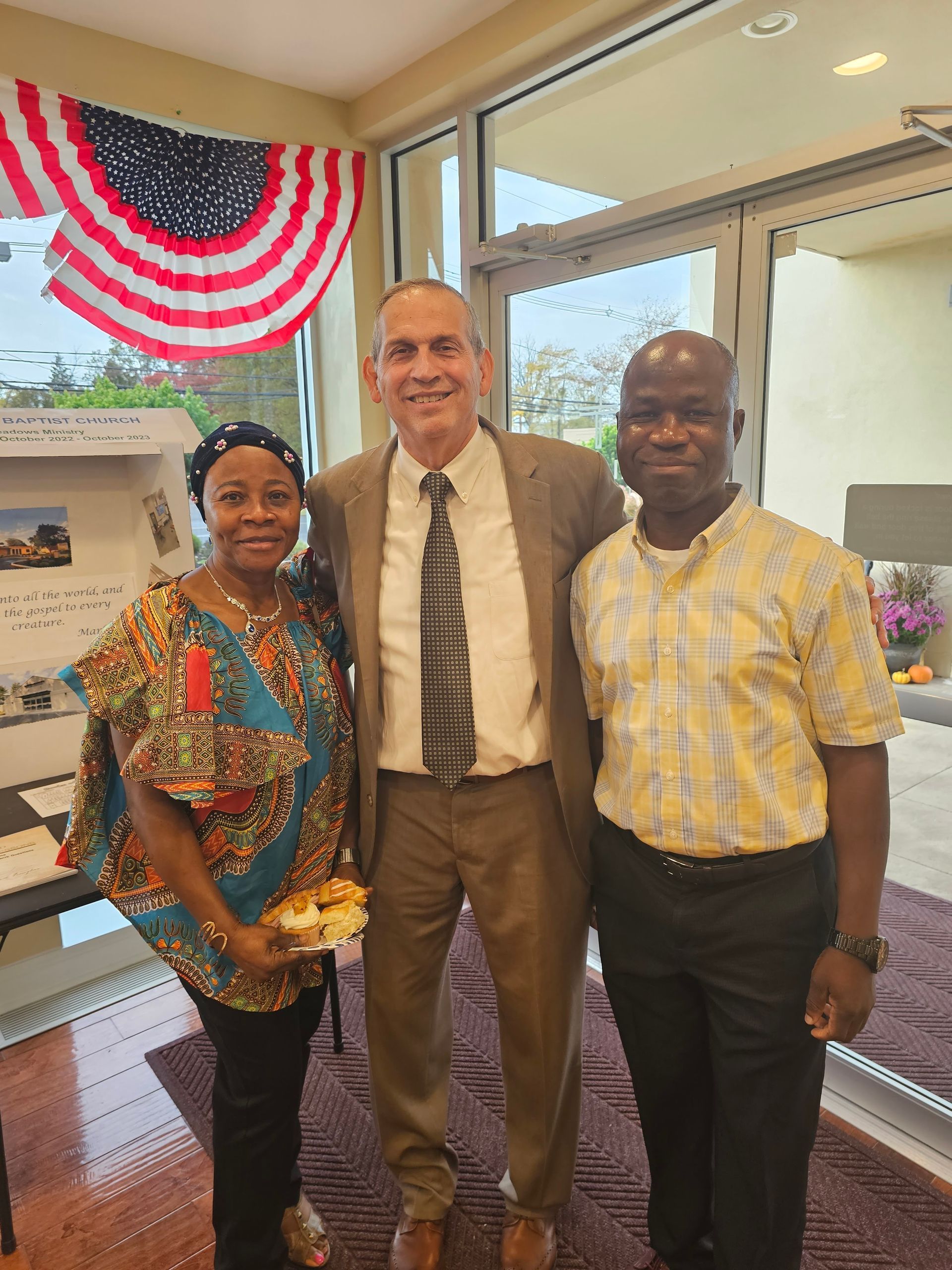Three people standing together indoors near a window, smiling, with red-white-blue bunting in the background.