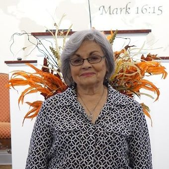 Smiling woman with gray hair and glasses, seated before orange floral arrangements and a Mark 16:15 wall sign