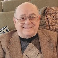 Smiling man in glasses and a tan blazer sitting on a couch