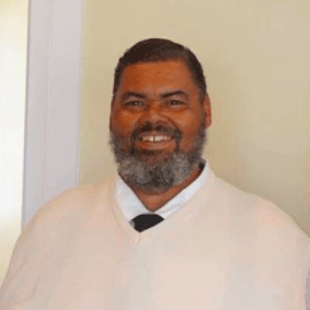 Smiling man in a white sweater and tie, standing against a plain light wall.