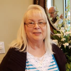 Woman with blonde hair and glasses standing indoors near a mirror and flowers
