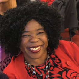 Smiling person in a red patterned top with a black curly wig, seated indoors.