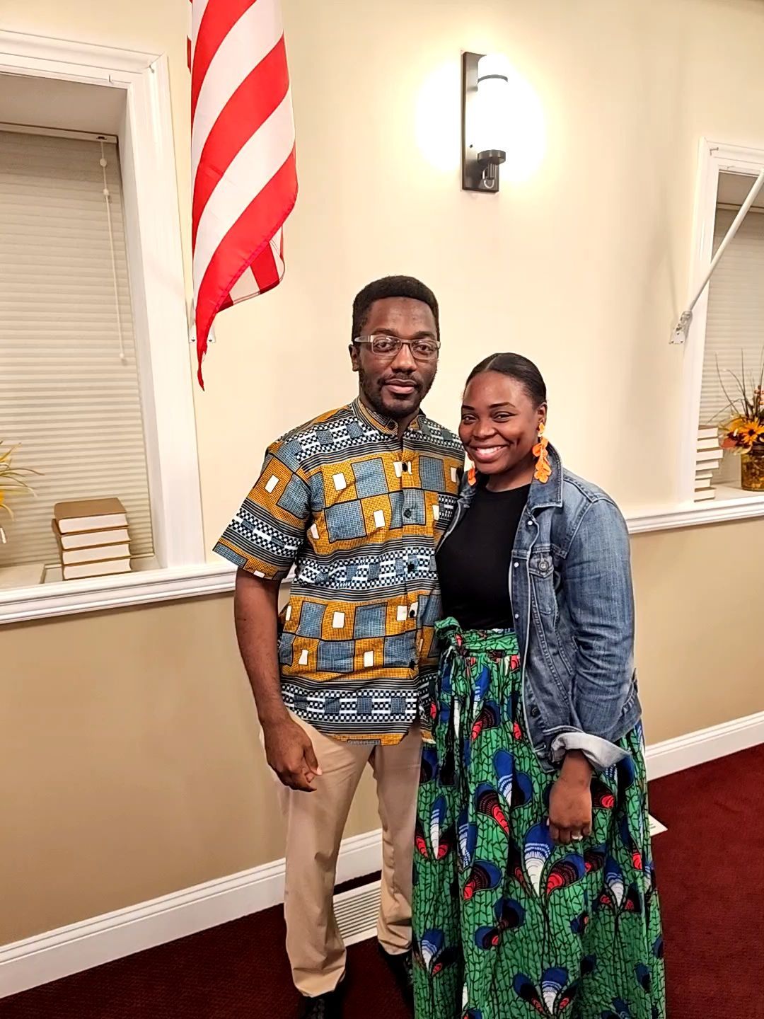 Two people smiling and posing indoors near an American flag and wall sconce.