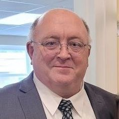 Smiling man in a suit and tie with glasses, standing indoors near a window.