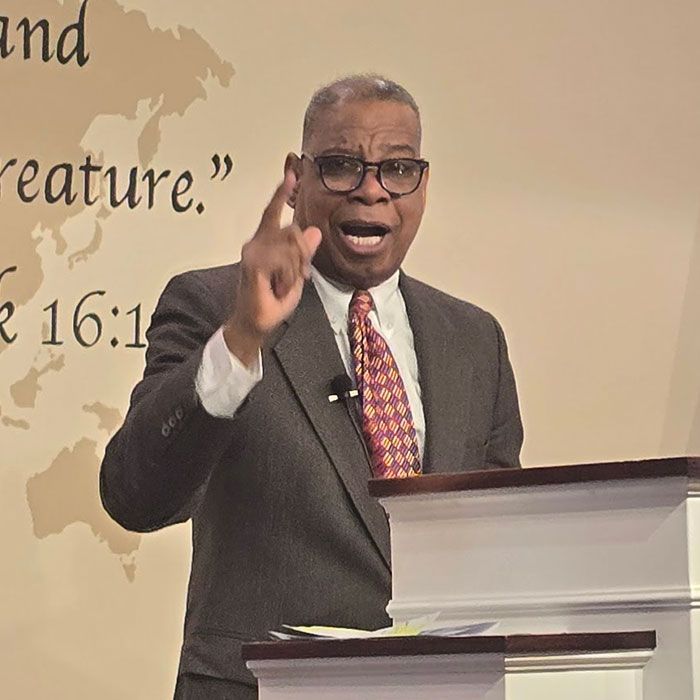 Man speaking at a podium, gesturing with one hand, in a church or conference setting.