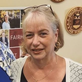 Smiling woman in a beige sweater and gold jewelry at an indoor event, with posters in the background