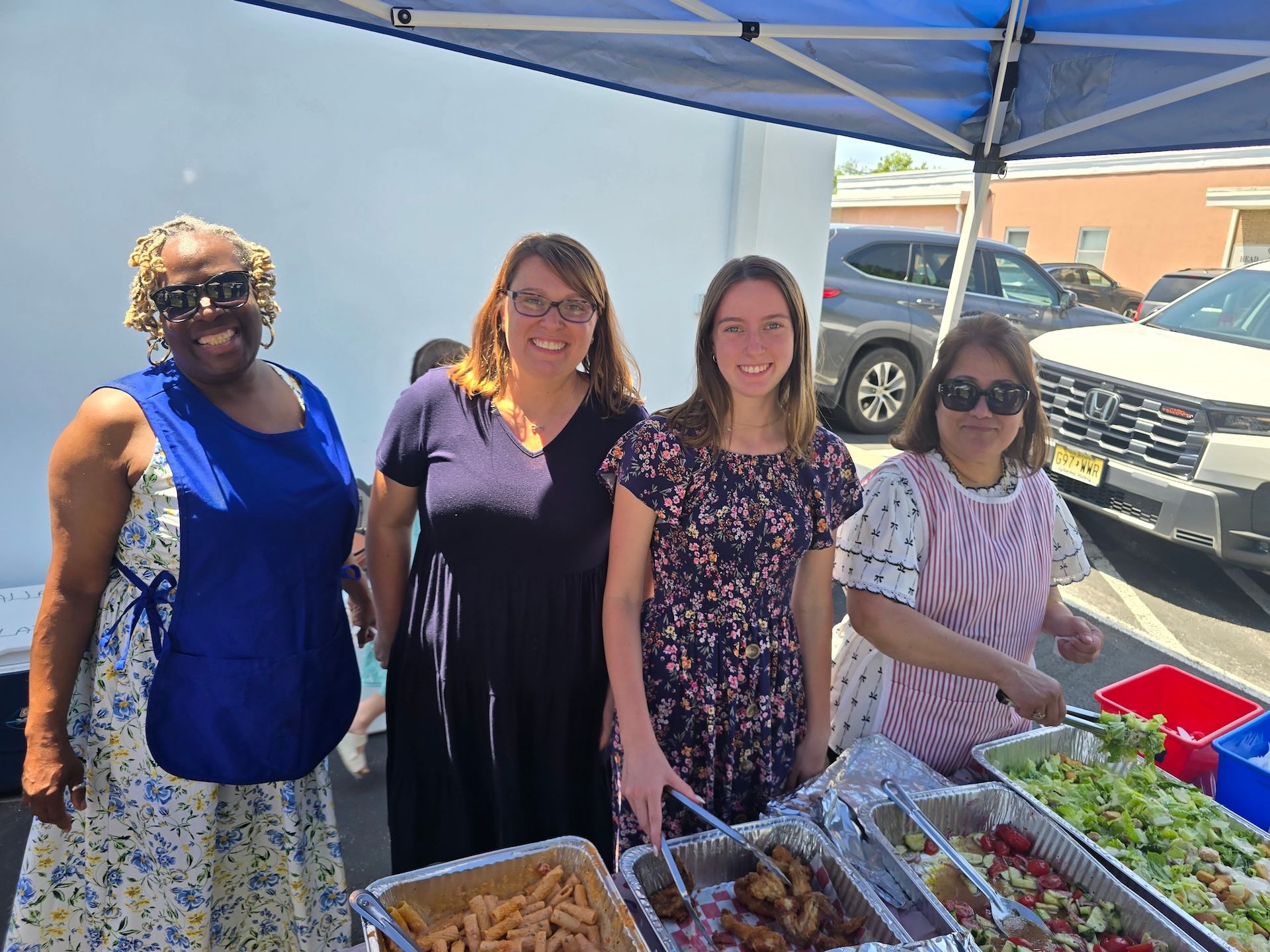 Four women stand behind trays of food at an outdoor event under a blue canopy.