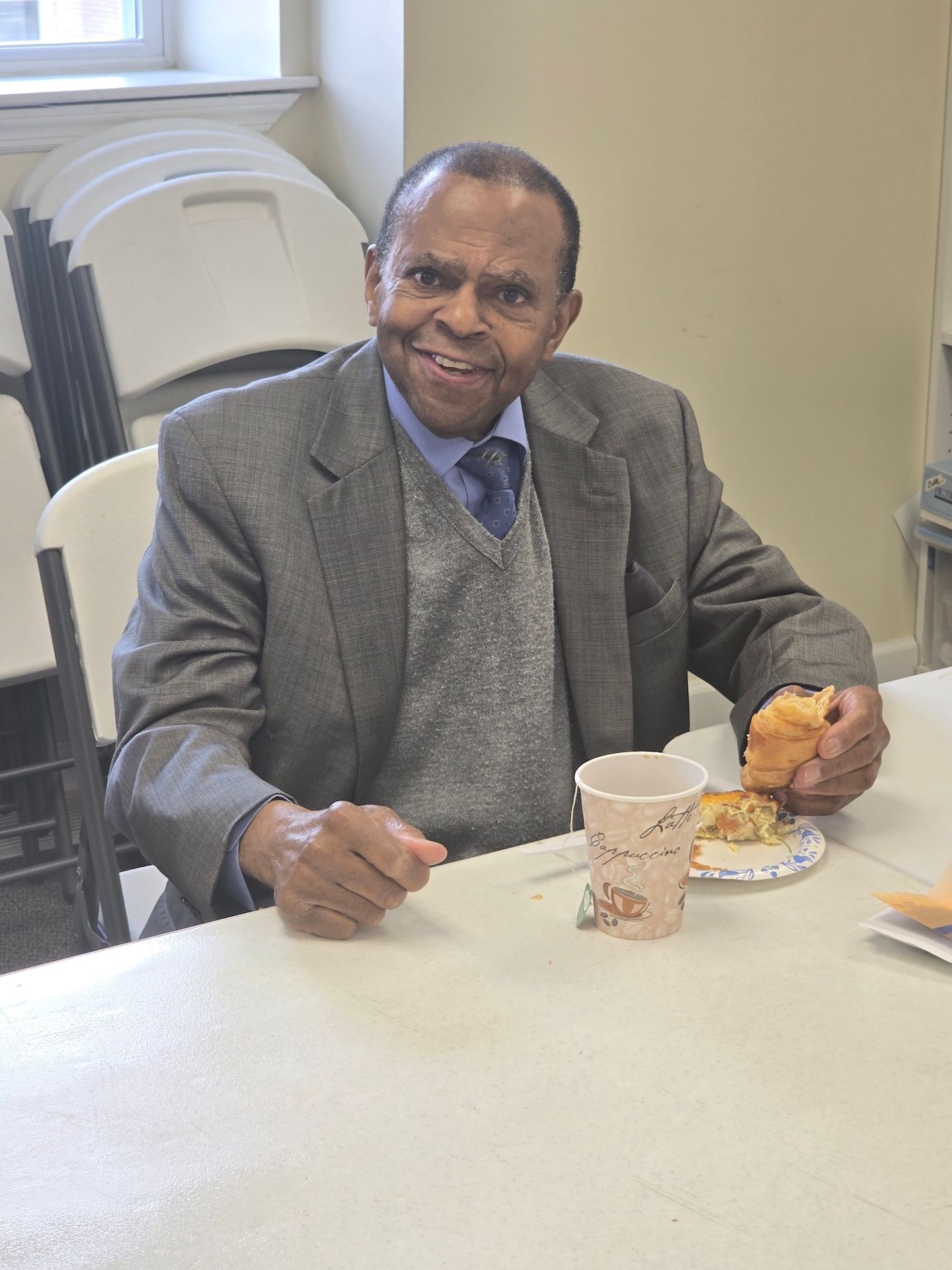 Man in a gray suit smiling at a table, holding a pastry with a cup of coffee beside him.