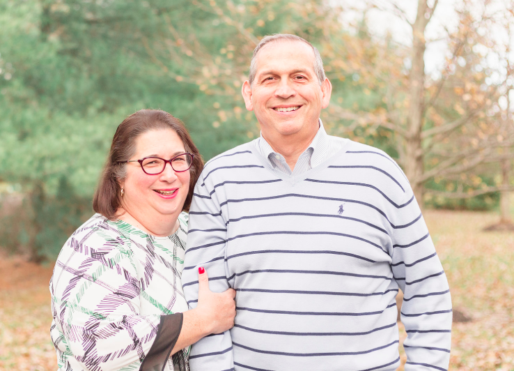 Smiling couple standing outdoors in a wooded area, wearing casual striped and patterned tops.