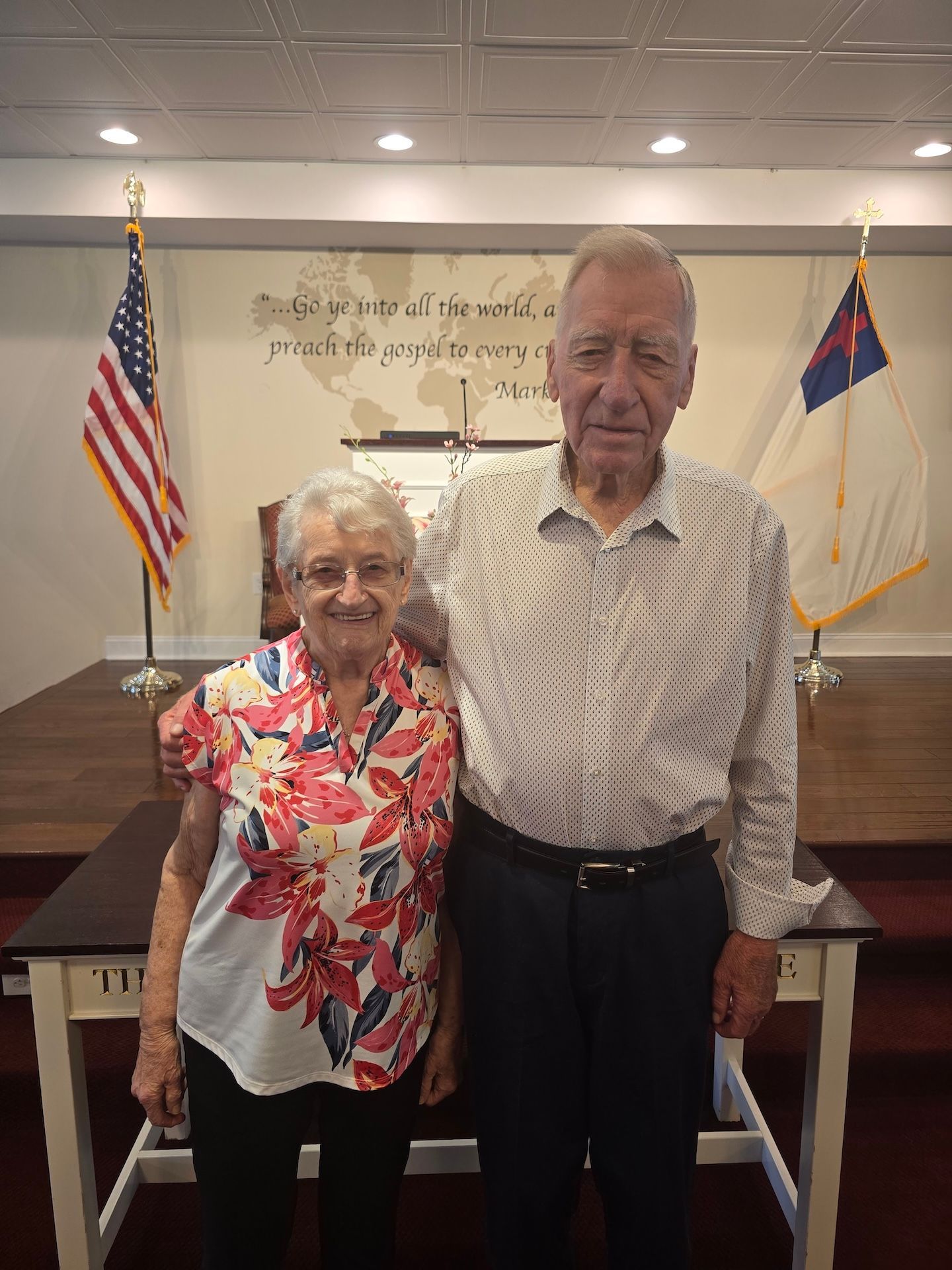 Two people standing indoors before flags and a wall sign, smiling for a photo