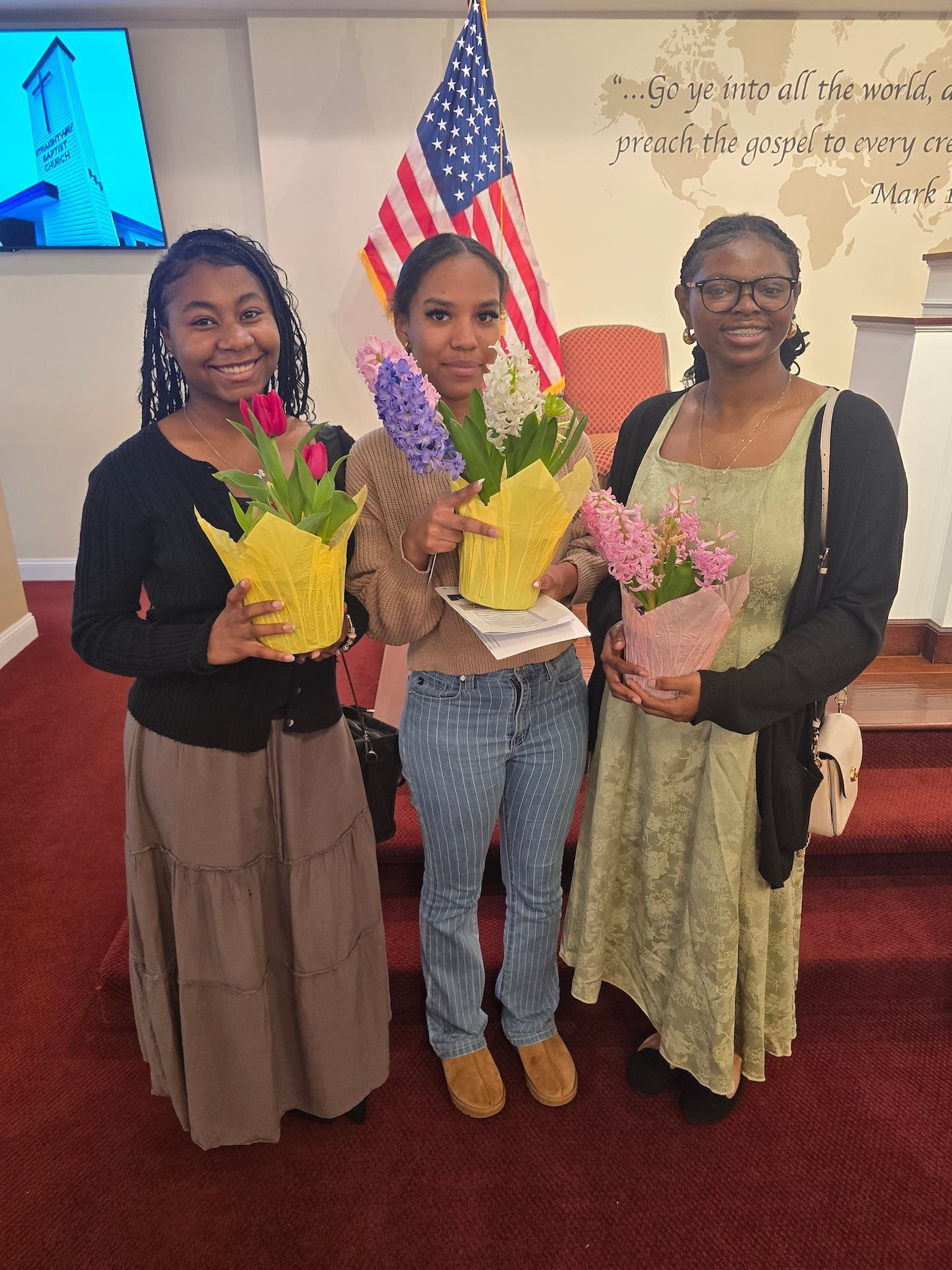 Three people smiling and holding bouquets in a decorated indoor room with an American flag.