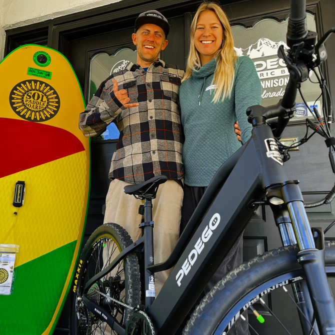 Two people pose with e-bikes beside a colorful surfboard display at a shop entrance