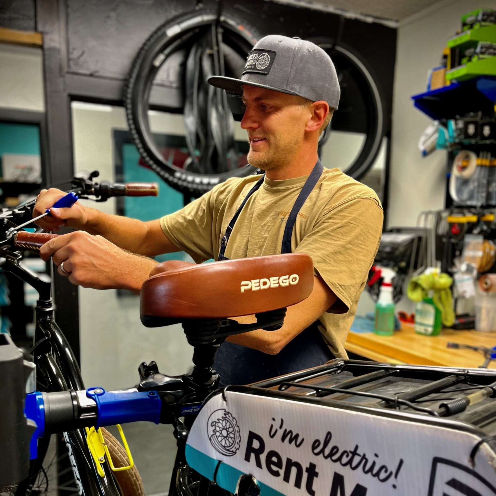 Man adjusting a brown bicycle seat in a workshop, with tools and an “I’m electric!” sign nearby
