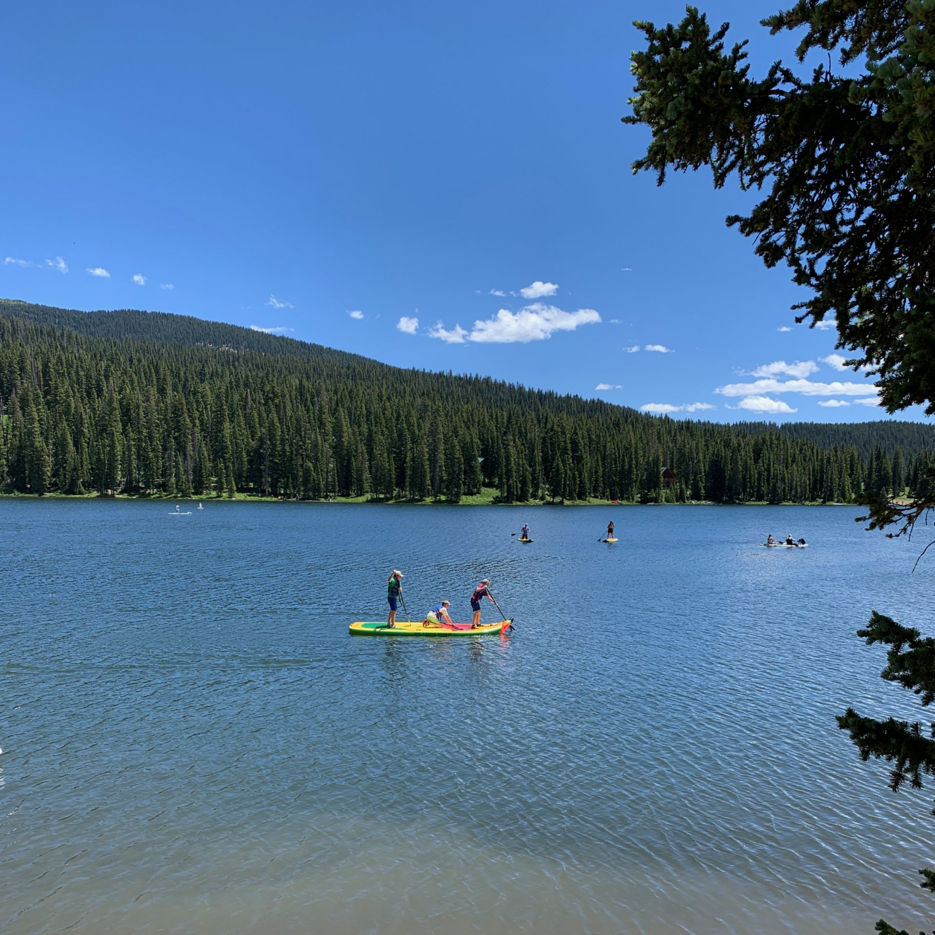 People paddle a colorful board on a calm blue lake with pine-covered hills under a clear sky.