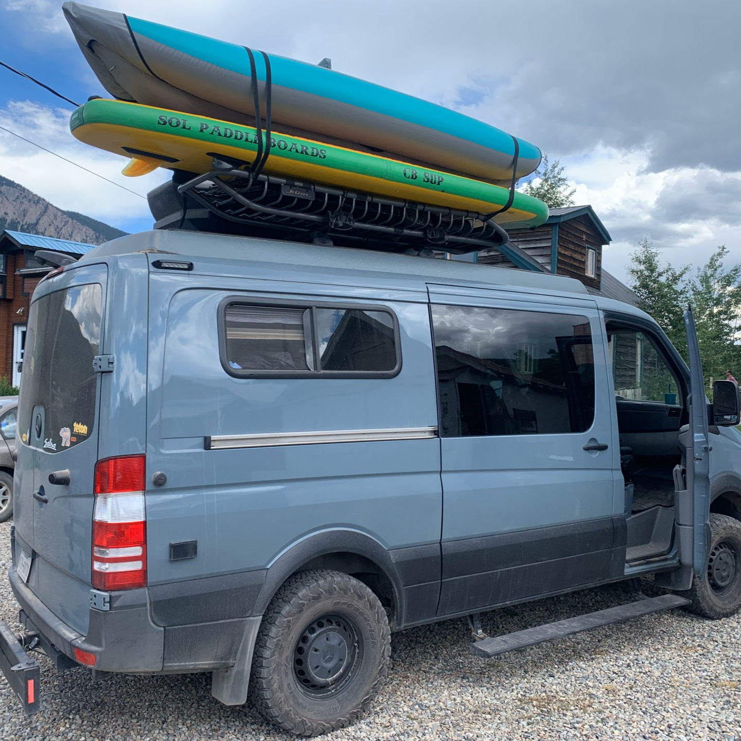 Gray van with two kayaks strapped to the roof, parked on gravel under cloudy skies.