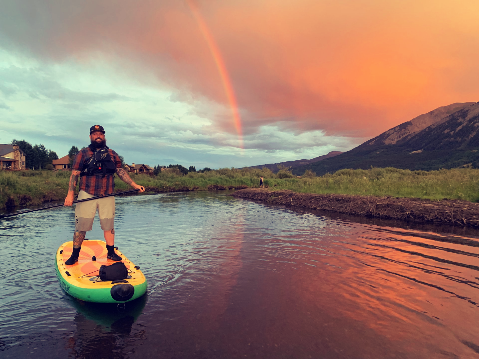 Person standing on a paddleboard on calm water at sunset, with a rainbow and mountains in the background