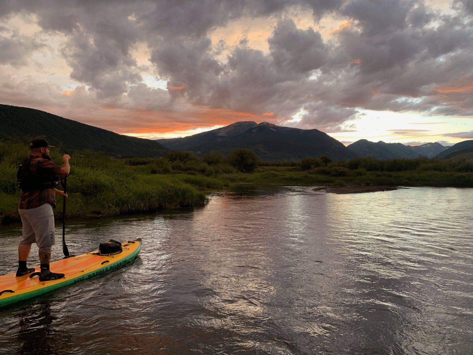 A person paddles on a yellow stand-up paddleboard in a river at sunset, with rolling hills in the background.