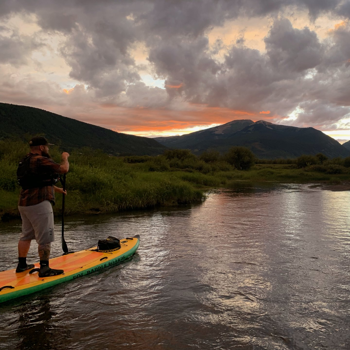 Person paddleboarding on a river at sunset with mountains and cloudy sky in the background