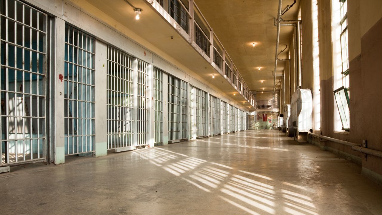 Empty prison corridor with barred cell doors and sunlight casting shadows on the floor