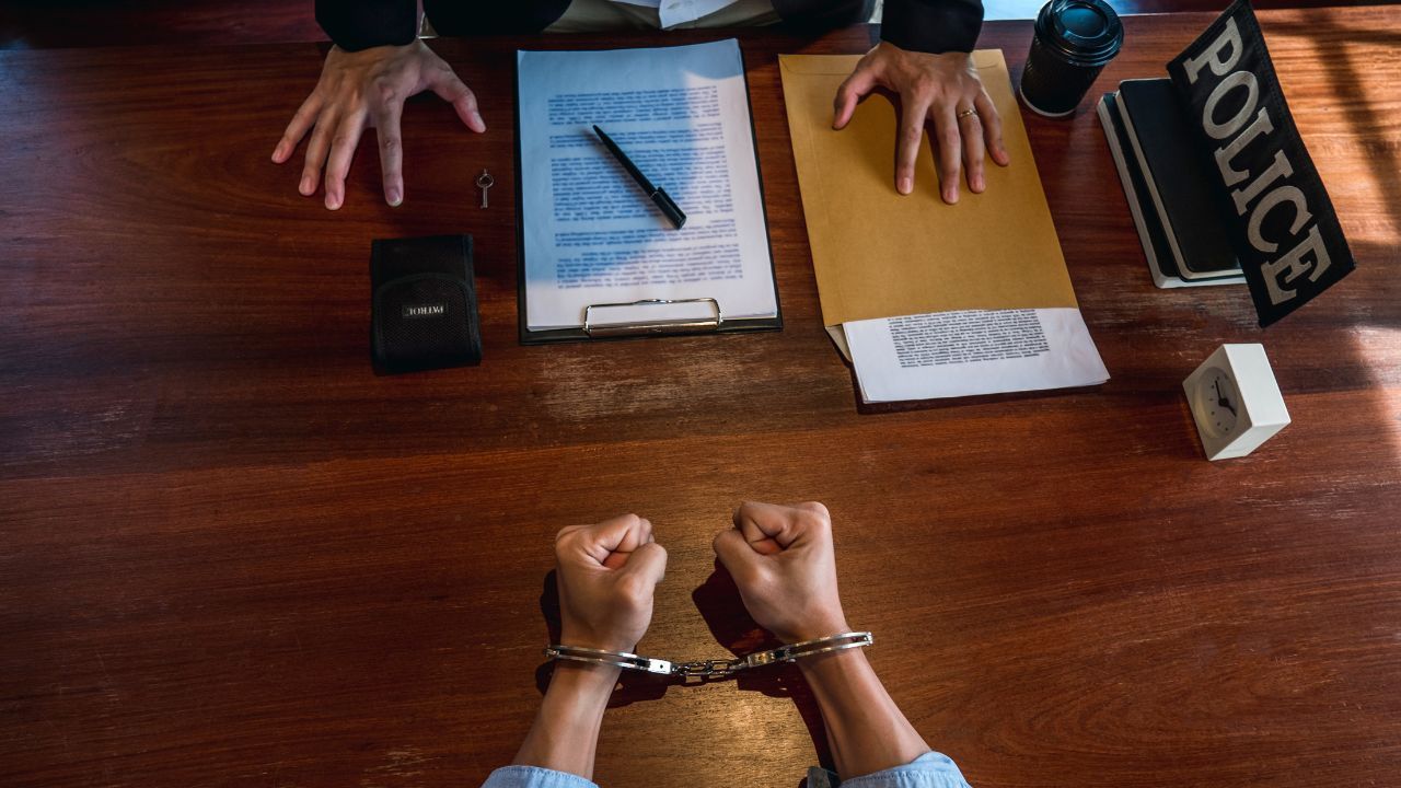 Handcuffed person at desk facing police officer’s hands, with case file, notebook, and POLICE sign.