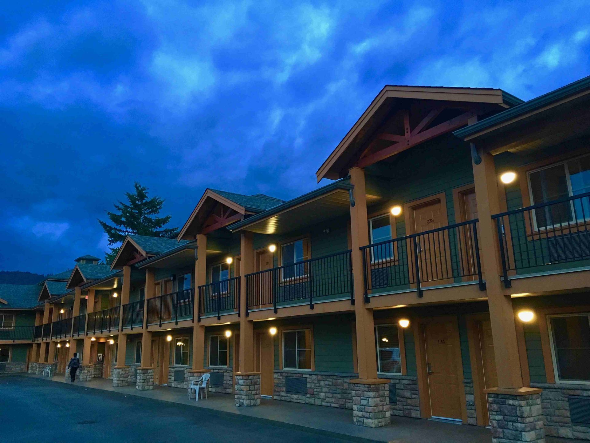 Motel exterior at dusk; teal walls, brown accents, balconies, and illuminated doorways under a stormy blue sky.