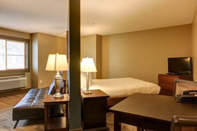Studio apartment with a bed, sofa, desk, and TV; earth-toned walls, wood floors.