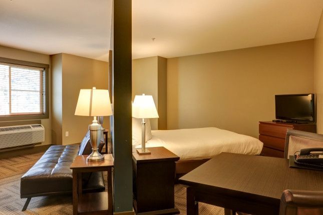 Studio apartment with a bed, sofa, desk, and TV. Brown tones dominate the decor.