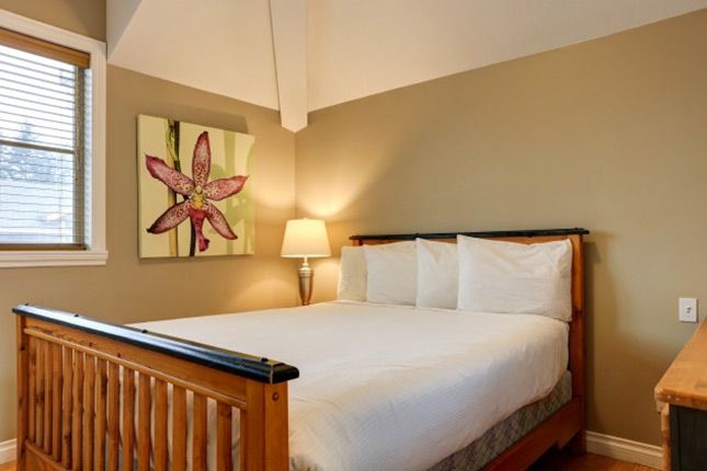 Bedroom with a wooden bed, white bedding, and a floral painting on the wall. Beige walls, window with blinds.