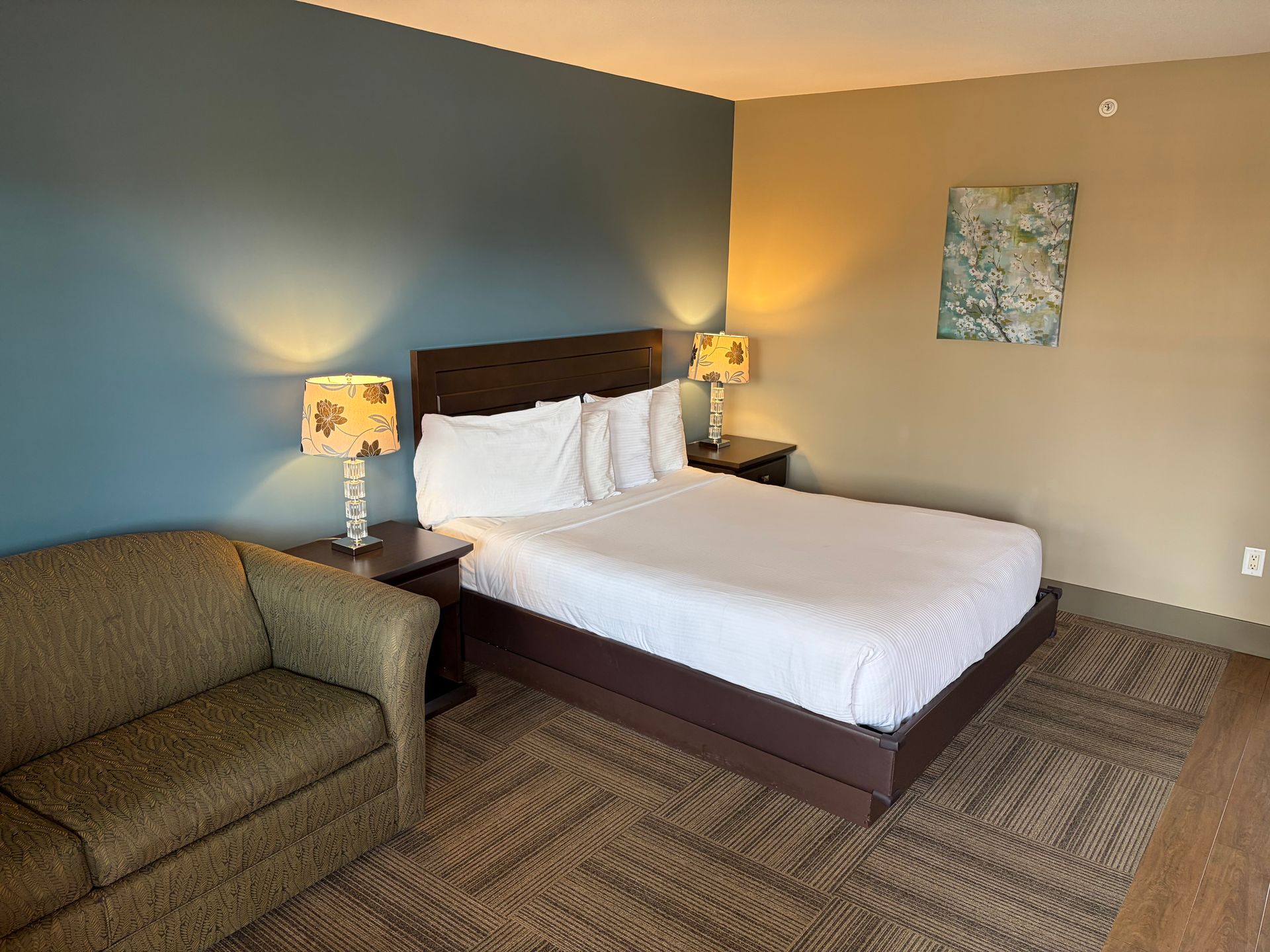 Hotel room with bed, armchair, nightstand, and lamps. Blue wall behind bed, painting on beige wall.