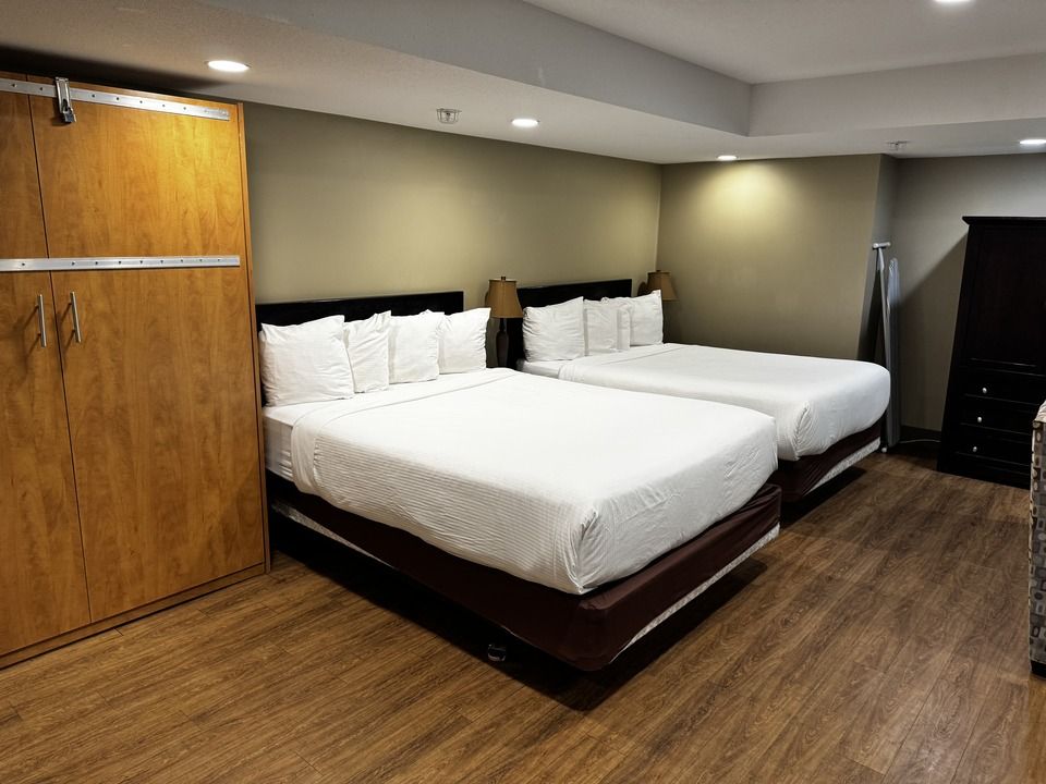 A hotel room with two beds, wooden floor, and a wardrobe. White bedding and neutral wall colors.