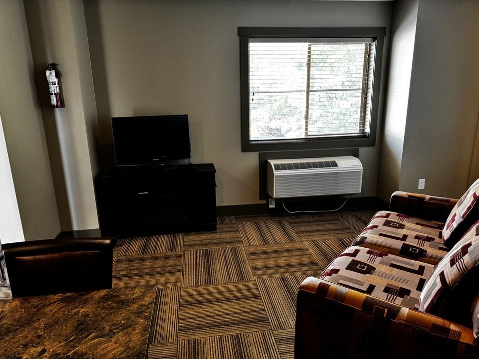Small hotel room with window, TV, couch, and air conditioner. Carpet has a wood-like pattern.
