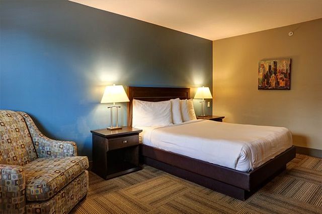 Hotel room with a bed, armchair, nightstand, and wall art; blue and tan walls, soft lighting.