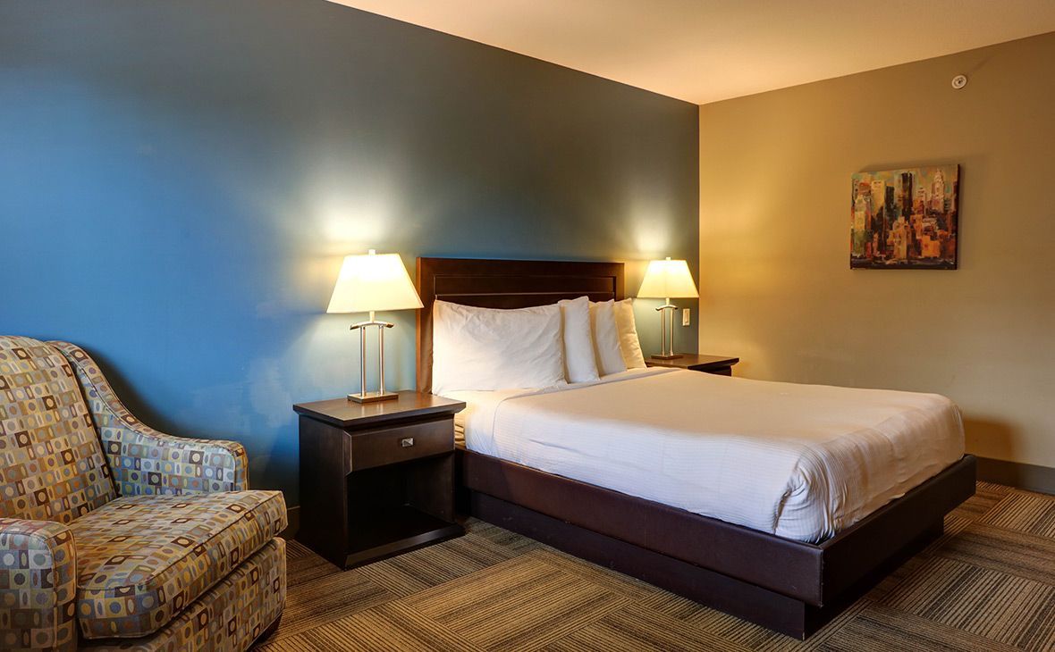 Hotel room with a queen bed, armchair, nightstand, and two lamps. Walls are blue and tan.
