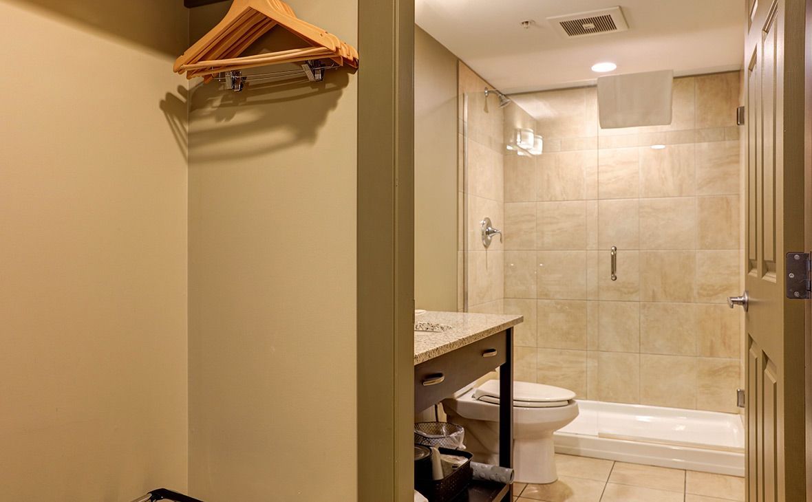 Bathroom and closet in a hotel room. Beige walls, shower, vanity, toilet, and hanging clothes hangers.