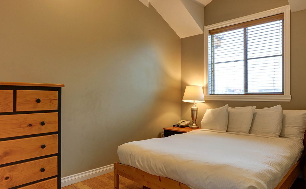 Bedroom with wooden furniture, bed, nightstand, lamp, and window with blinds. Brown walls, light wood floor.