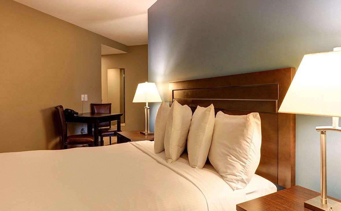 Hotel room with a bed, lamps, a table, and a hallway. Beige walls, teal accent wall, and wooden furniture.