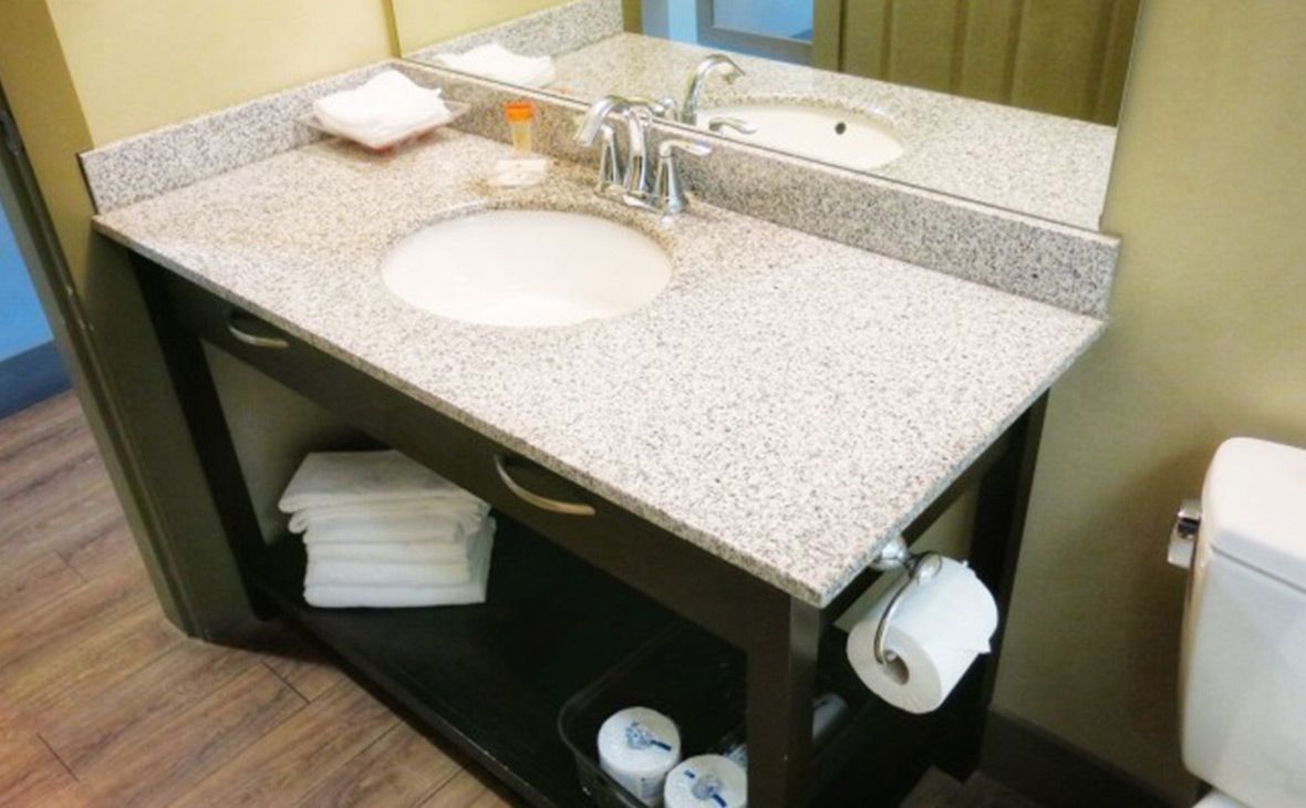Bathroom sink with granite countertop, mirror, and toilet paper. Stacked towels and toiletries are visible.