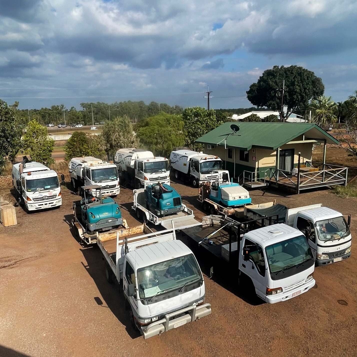 A fleet of utility trucks parked on dirt in front of a small building under a cloudy sky — Industrial Power Sweeping Services Pty Ltd In Knuckey Lagoon, NT