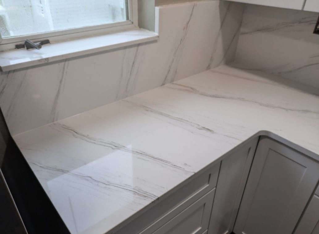 White marble-look kitchen countertop and backsplash with gray cabinets under a window