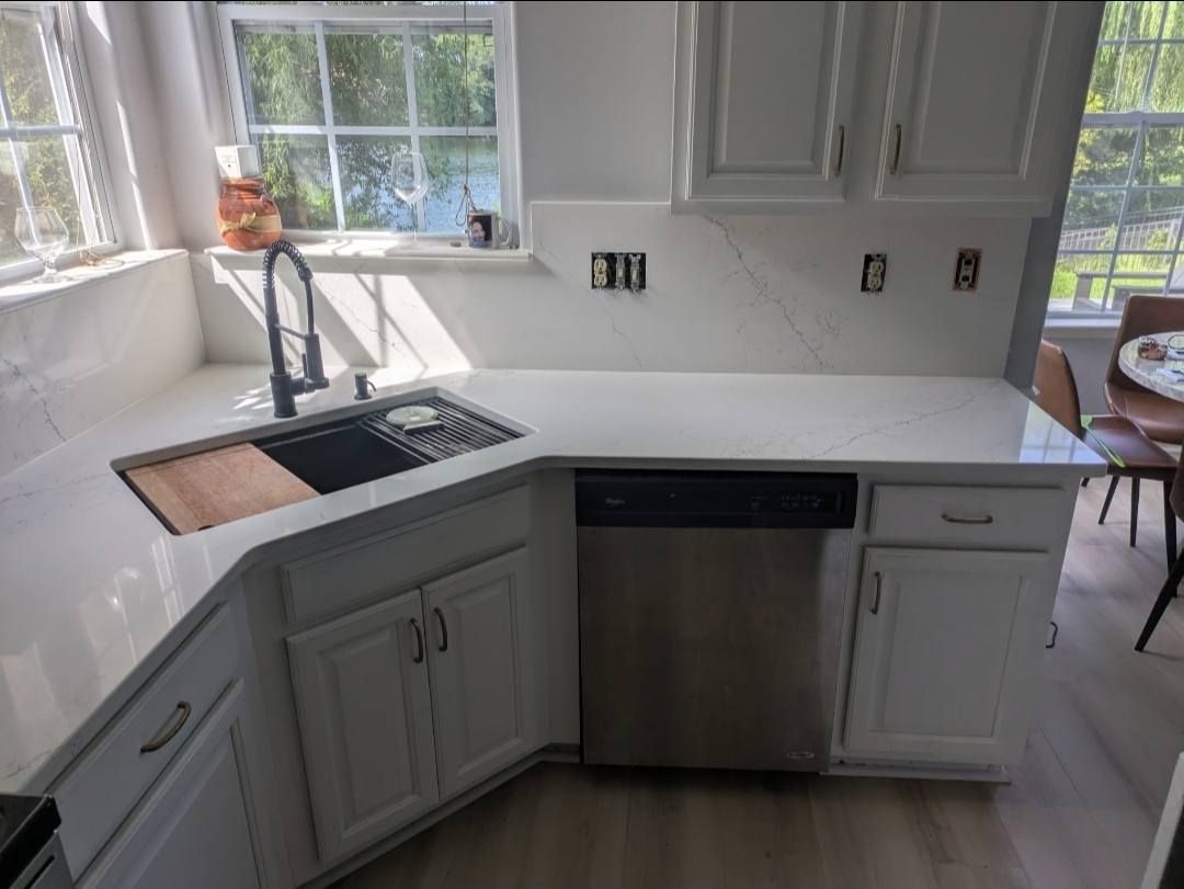 Bright white kitchen with a stainless dishwasher, corner sink, and large windows letting in sunlight
