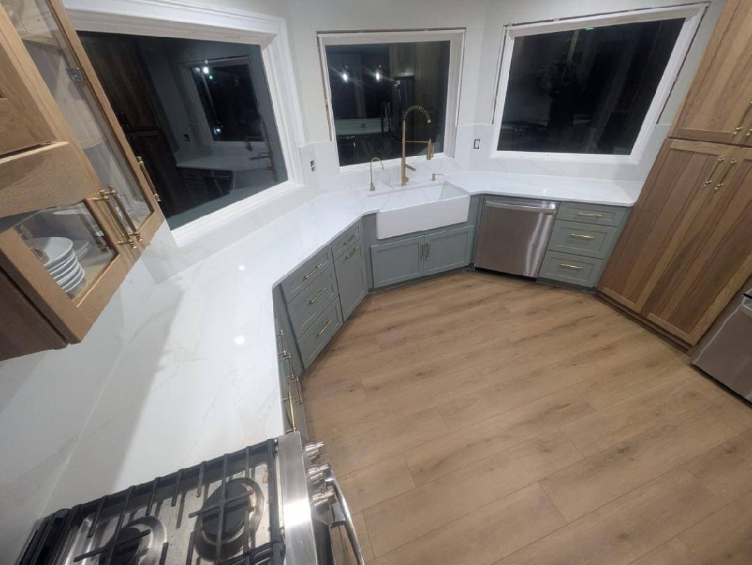 Modern kitchen with white countertops, gray cabinets, wood flooring, and windows over the sink.