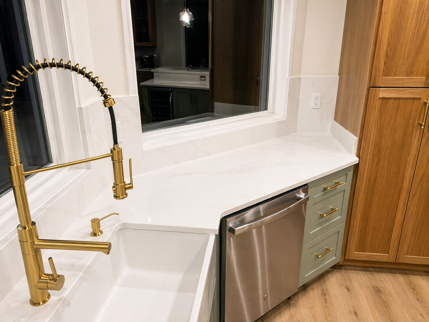 Modern kitchen sink with brass faucet, white countertops, stainless dishwasher, and wooden cabinets