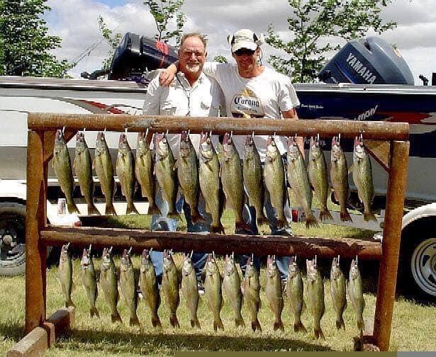 Two men standing with a wooden rack displaying numerous fish, next to boats, outdoors.