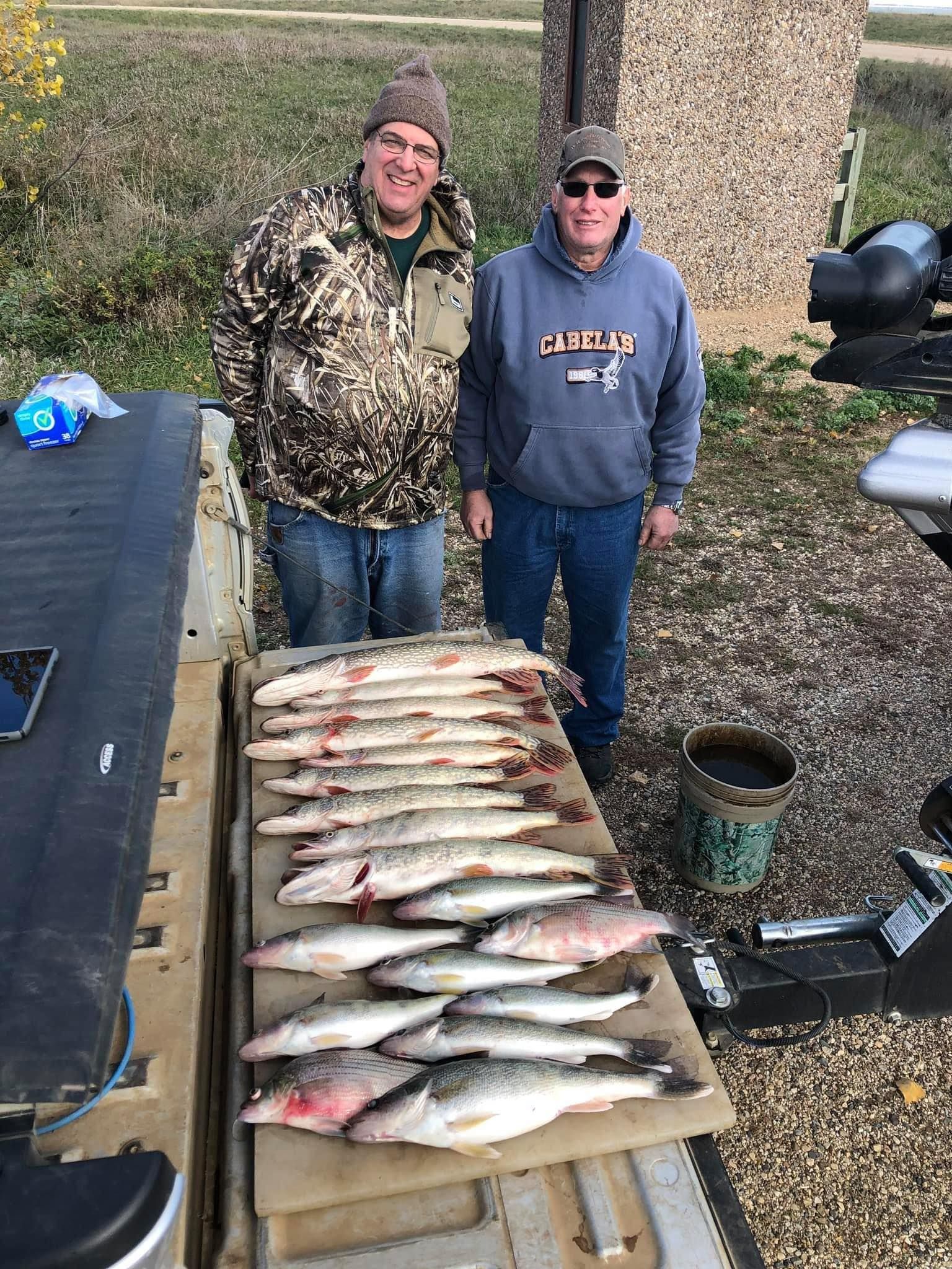 Two men stand behind a row of caught fish. They are outside with a neutral background, and one is wearing camo.