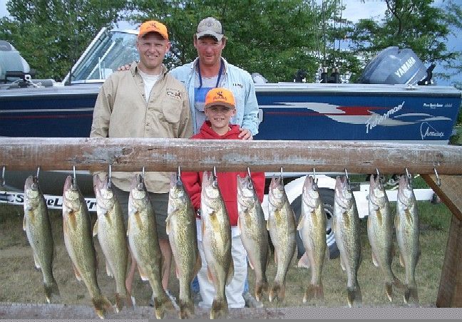 Three people posing with a row of caught fish. Blue boat in background.
