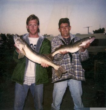 Two men holding up large fish; outdoor setting, trailer in background.