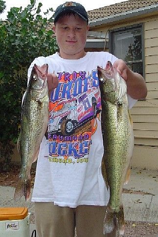 Boy holding two large fish, smiling. Outdoors, wearing a hat and graphic t-shirt.