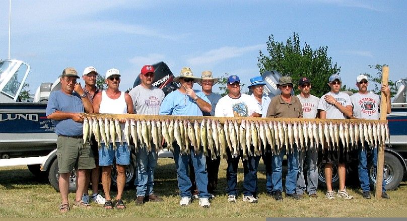 Group of people with a wooden rack holding many fish; boats and a tree are in the background.