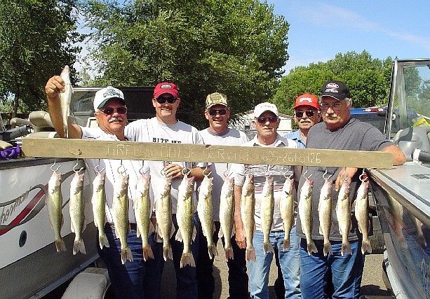 Six people holding a board with many caught fish on a boat, sunny day.