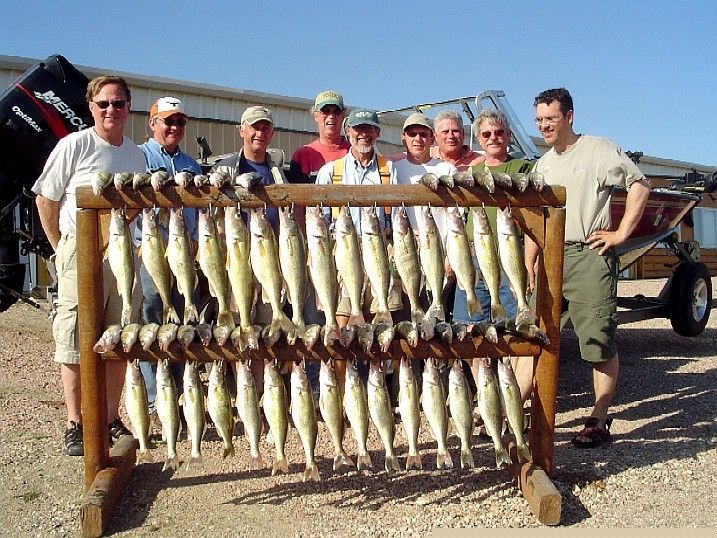 Group of people with a wooden rack of caught fish in front of a boat. Outdoors, sunny.